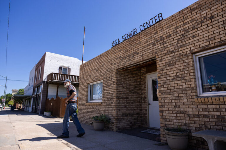 A man with a white beard is shown leaving a one-story brick building on a small town street. The building has a sign that reads,