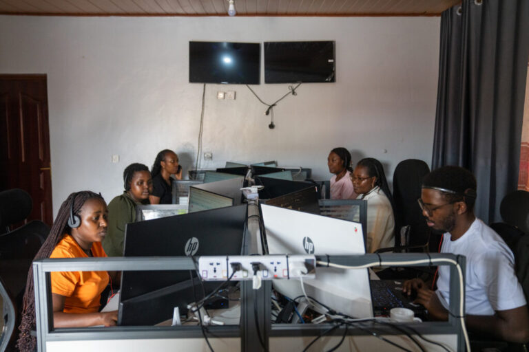 The call center in Muhanga where Zipline employees take calls and organize supplies to be sent via drone to people who need them. Six people are sitting at small desk, looking at computer monitors and wearing headsets.