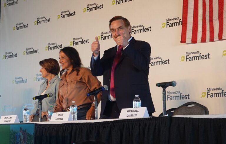 Mike Lindell stands on stage in a dark suit and red tie and gives two thumbs up. Next to him are Sen. Amy Klobuchar on the left and Lisa Demuth, speaker of the state house, in the middle.