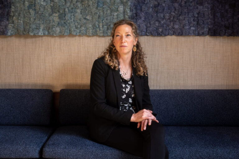 Noella Sudbury is the founder of Rasa Legal, a non-profit that uses technology to help people expunge their criminal records. She is sitting on a couch with her hands in her lap. Sudbury has long, curly hair and she is wearing a dark suit and a dark floral top.