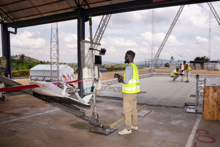 One drone is prepared for take-off while another is collected from the landing mechanism. A technician, wearing a yellow safety vest, looks at a laptop while preparing the white and red drone for launch. Two other technicians are holding another drone.