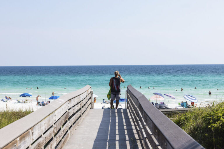 A man wearing shorts and a sleeveless top stands on the wooden boardwalk that leads to the sand at Ed Walline Regional Beach Access in Santa Rosa Beach, Florida, on August 25. Blue waters are in the background.