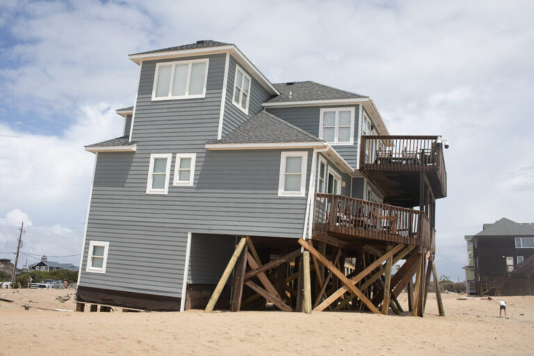 A three-level house with gray siding is tilted askew as one side of it, supported by wood stilts underneath, sinks into the sand.
