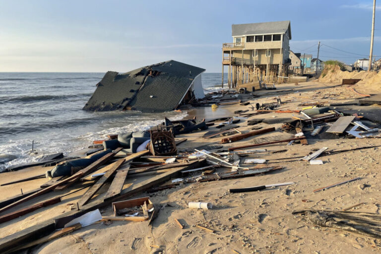 A collapsed house lies in shallow ocean water on the coast. Debris such as planks of wood and a sofa lie scattered on the sand. In the background is a two-story house that sits on top of tall wooden stilts.