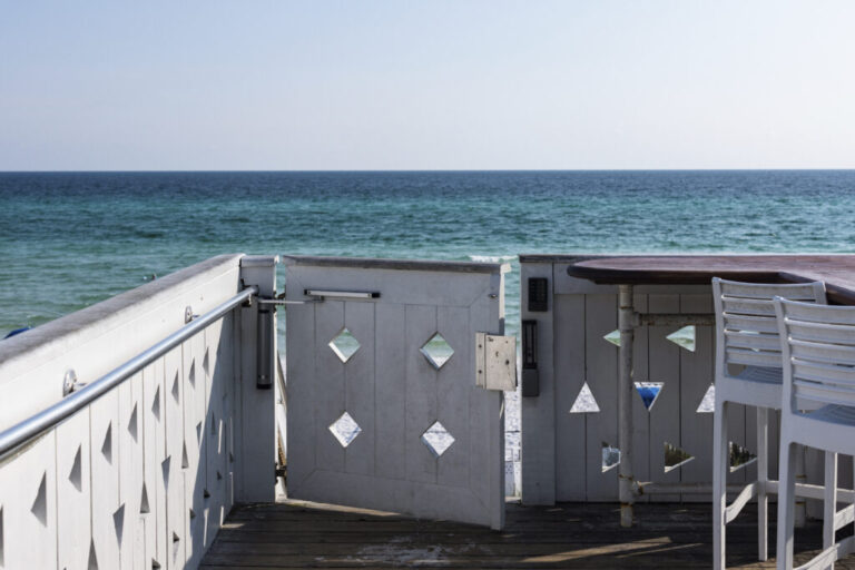 A gray wooden gate on the edge of a wooden beachside deck is equipped with a keypad to grant access to the beach behind it, near the Coleman Pavilion obelisk in Seaside, Florida. Blue waters are in the background.