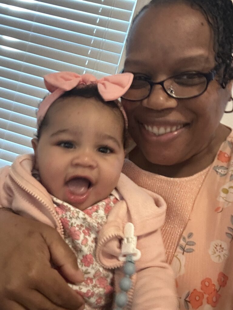 Yalanda Lattimore, 59, holds her 1-year-old granddaughter, Mavee. Lattimore is smiling, and Mavee has her mouth wide open. Mavee is dressed in pink, and around her head she's wearing a pink headband with a bow.