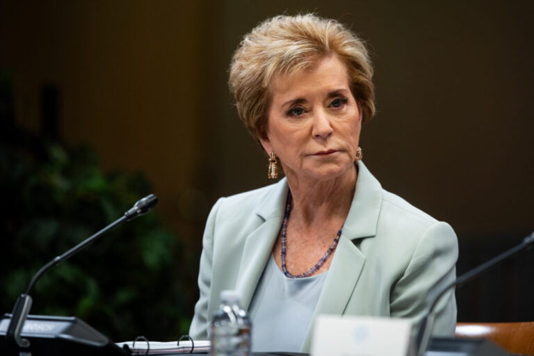 An older woman with blonde hair and a powder blue suit sits at a table with a microphone in front of her.