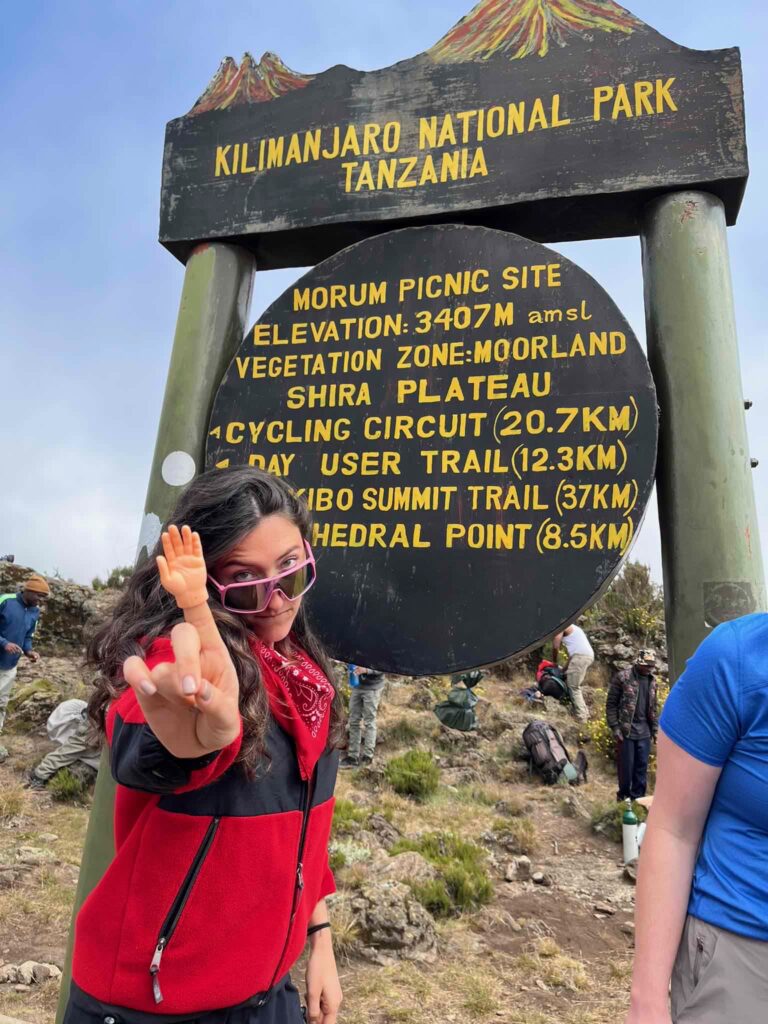 A vibrant young woman is shown in front of a sign for Kilimanjaro National Park, Tanzania. She is looking over her sunglasses and gesturing at the camera with a finger puppet on her index finger.
