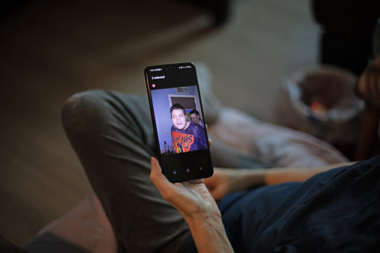 Debbie Willson holds her smartphone, which is displaying a photo of her son, Adam Willson, who died at age 27 in 2024. Adam is wearing a T-shirt that says