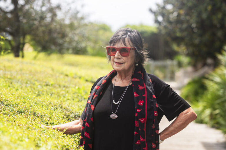 Linda Miller stands outdoors, surrounded by foliage, at the Western Green outdoor venue in Rosemary Beach on August 20. She's dressed in black and is wearing red sunglasses.
