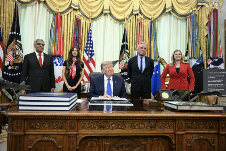 President Donald Trump sits at the Resolute Desk with several people behind him.