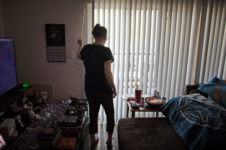 Debbie Willson twists a rod to close long vertical shades in her dimly lit home. Her back is to the camera.