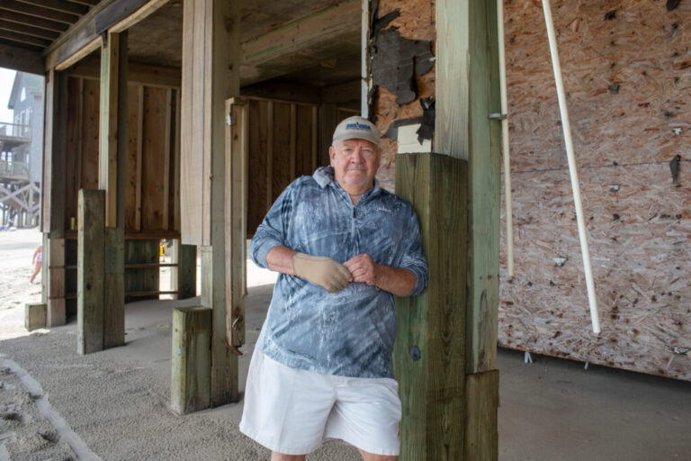 Bill King is standing by a house with exposed plywood and wooden beams. He's wearing white shorts, a gray shirt and a beige visor.