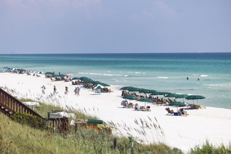 Beachgoers lounge on wooden reclining beach chairs, with green umbrellas providing shade, in Rosemary Beach, Florida, on August 25. The sand is white, and the water is blue.