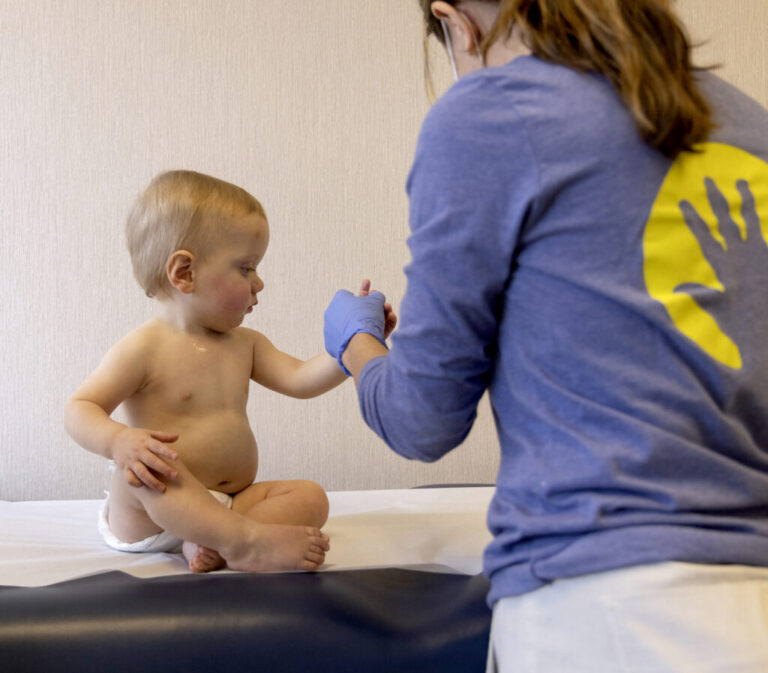A child, who appears to be about a year old, is sitting on an exam table, wearing a diaper. A medical professional is holding the child by the hand.