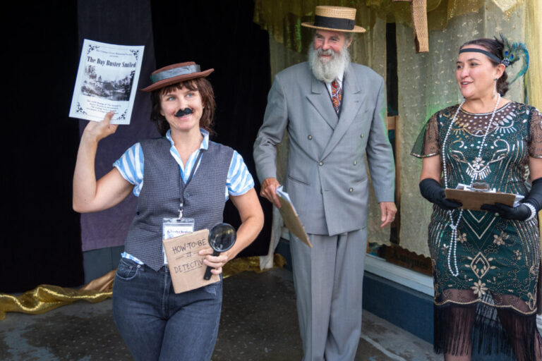 Liz Bartholomew (left) wins for best Buster Keaton look-alike after being awarded by Dana Merryday (center) and Jennifer Ferraez Chartrand during a 1920s themed costume contest. Liz is wearing a porkpie hat, a fake mustache, a vest over a striped short-sleeve button down shirt and carrying magnifying glass and a handwritten sign that says
