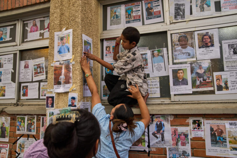 A young boy is helped by a women to put up a photograph of her missing relative on a wall alongside photographs of missing and deceased people displayed outside a government hospital in Kathmandu, Nepal, Sunday, Aug. 30, 2026.