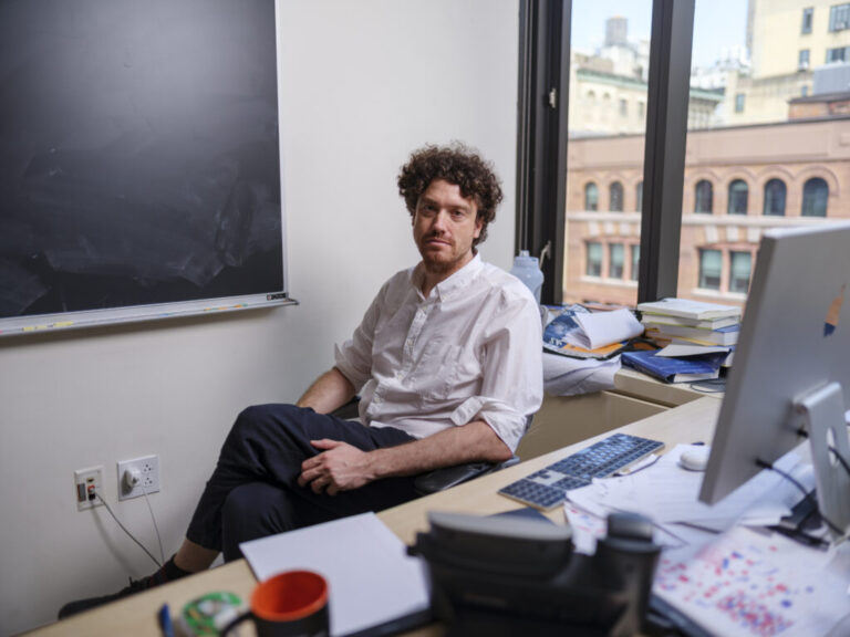 Tristan Buckmaster, mathematician at New York University, poses for a portrait in his office in Manhattan. He is wearing a white button down shirt and dark slacks. Behind him is a blackboard and a window. In front of him is a desk with a computer, keyboard and some papers.