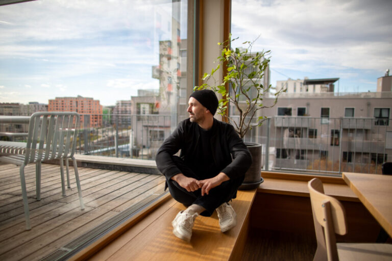 Sebastian Schublach sits next to the windows of his buildings shared kitchen and hosting space. Schublach has lived in this climate-friendly affordable apartment in Vienna for six years with his wife and two kids. At top: Schlubach sits in the building’s light filled rooftop kitchen and dining room.