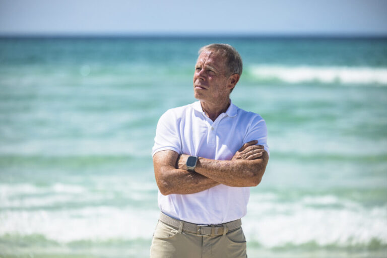 Dave Rauschkolb stands on a beach off Scenic Highway 30A on August 20. He's wearing a white polo shirt and khaki pants, and his arms are crossed across his chest. Water and waves are behind him.