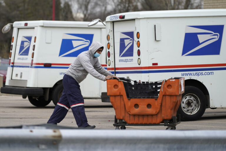 A U.S. Postal Service employee wearing dark blue pants, a gray hoodie and a light blue medical mask pushes a large orange plastic cart in a parking area outside a post office in Wheeling, Illinois, in 2021. Two Postal Service delivery trucks are in the background.