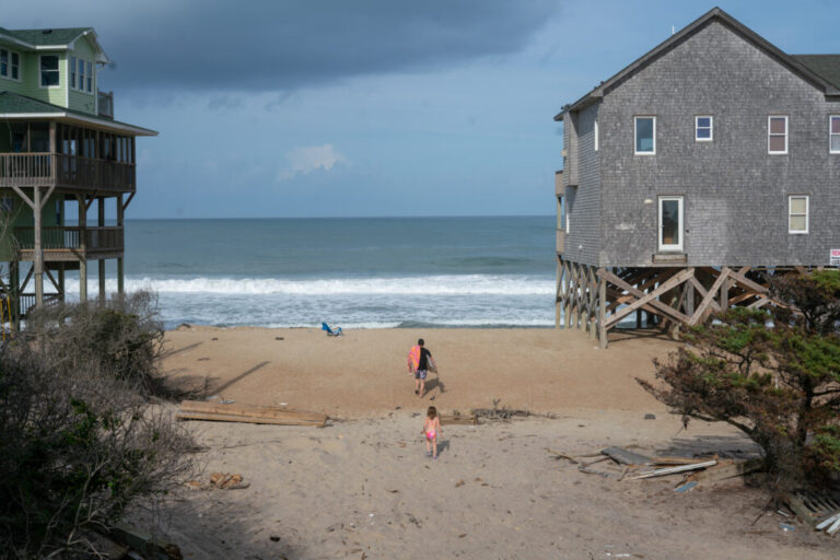 A man and girl clad in swimwear, their backs to the camera, walk cross the beach, toward the ocean. They are walking between two large multilevel houses that are perched on wooden stilts.