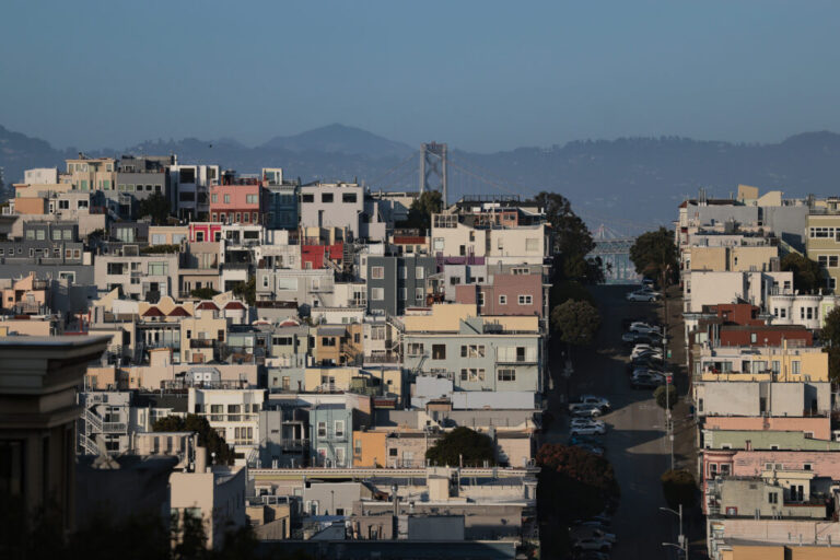 This photo taken at a distance shows houses and buildings in San Francisco's North Beach neighborhood. The structures are mainly various pastel colors such as pink, light blue and butter yellow. Mountains rise in the background.