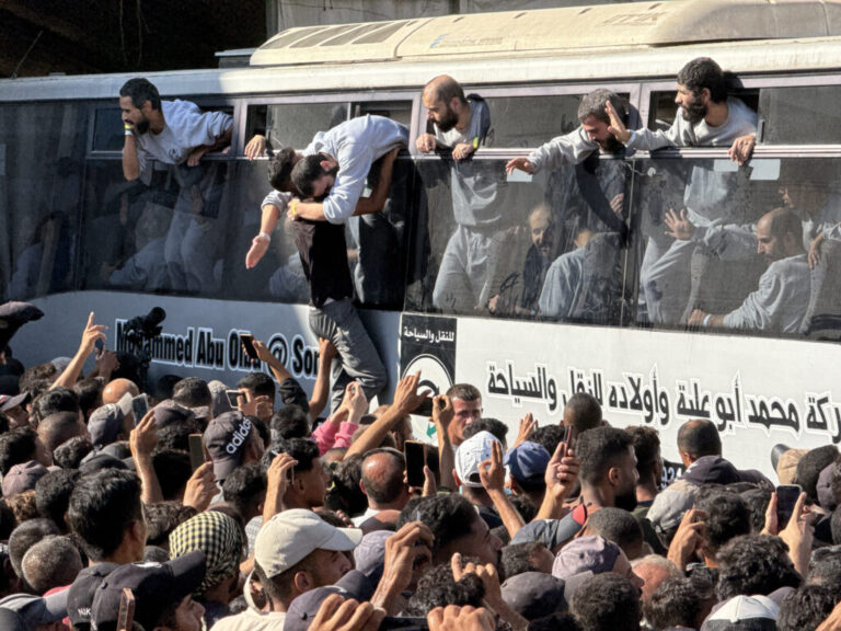 Family members of freed Palestinian prisoners crowd around a bus that's filled with the prisoners. Some of the prisoners are peering their heads out the windows of the bus.