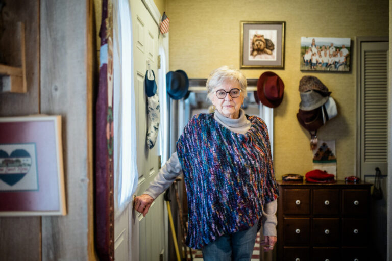 Suzann Roberts, 90, stands in her home in Ann Arbor, Michigan. She has short gray hair and is wearing glasses. She has a colorful poncho over a gray sweater.