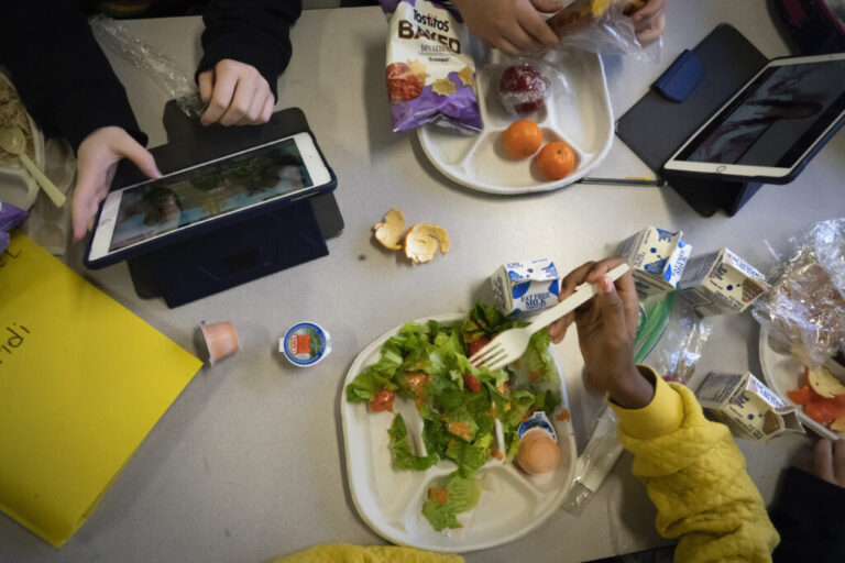 Photographed from above, seventh-graders sit together at a cafeteria table during lunch at a school in Brooklyn, New York, on February 10, 2023. One student is eating a salad, and another has a plate with tortilla chips and fruit on it. Two tablet devices and multiple cartons of milk are also on the table.