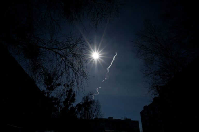 A white rocket trail appears against a dark blue sky over Irpin on March 12, 2022. Tree branches are silhouetted against the dark sky.