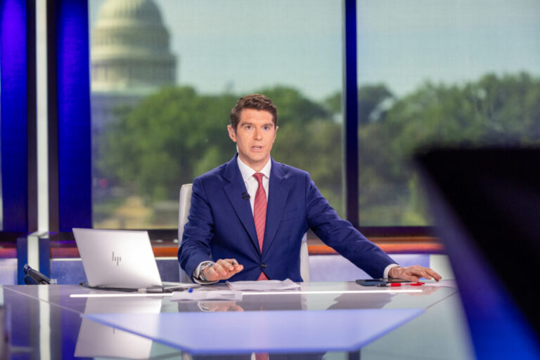 Wearing a blue suit, Benjamin Hall sits behind a news anchor's desk as he hosts