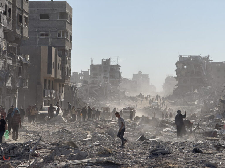 Palestinians walk amid rubble, debris and the skeletons of heavily damaged multistory buildings.