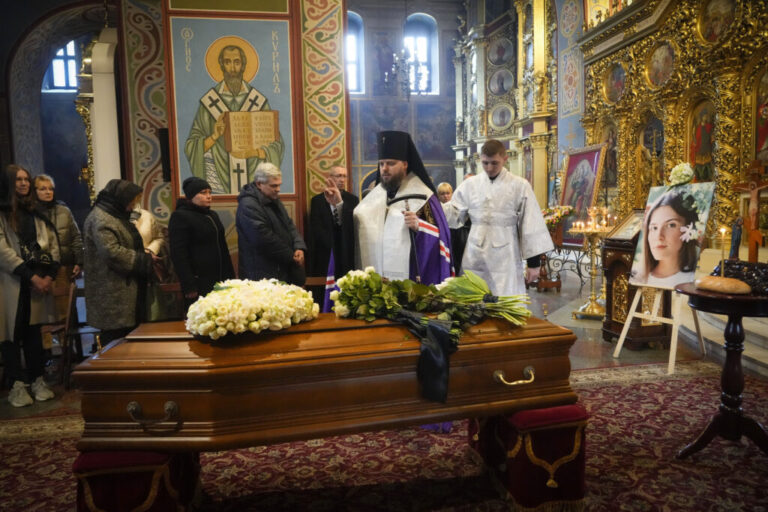 A priest wearing vestments prays over Oleksandra Kuvshynova's brown wooden coffin during a funeral service inside St. Michael's Cathedral in Kyiv on March 20, 2022. The coffin has white flowers placed on top of it.