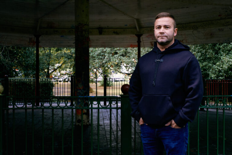 Shane Thomson stands outdoors. He's wearing blue jeans and a dark blue hoodie.