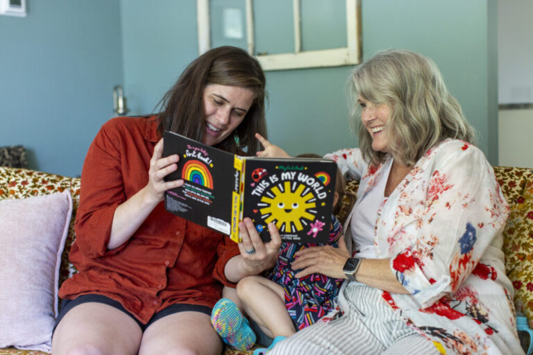 Stacia Bourne; her mother, Mary Jo Galusha; and Bourne's child are sitting on a sofa at Galusha’s home in Newark, Delaware. The child is sitting between the two women, and Bourne is holding up a children's book as the three enjoy it together.