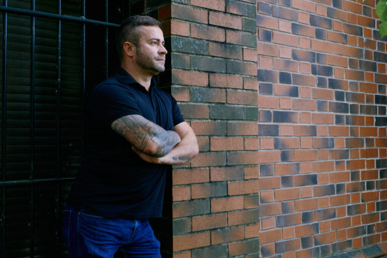 Shane Thomson stands next to a brick wall. He's wearing jeans and a navy blue short-sleeved shirt.
