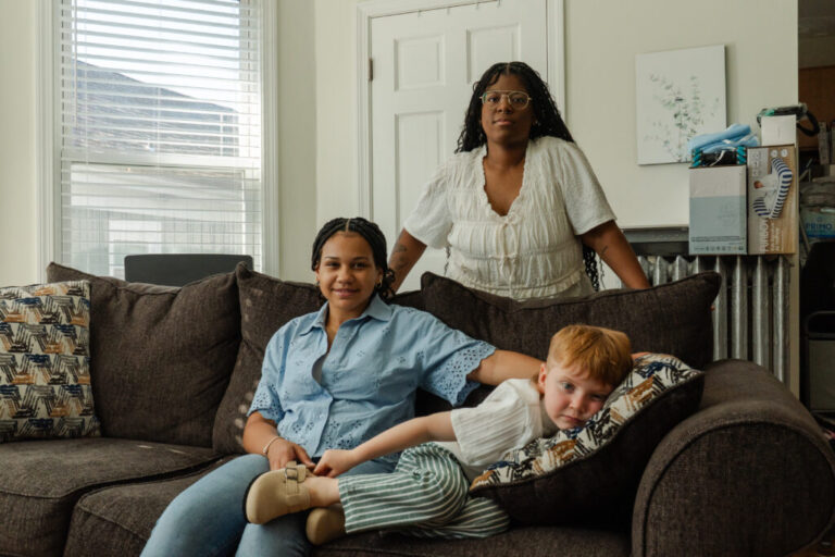 Emani Pike, 14, is sitting on a couch with Austin Pike, 4, who is laying down. Marissa stand behind them. Emani has long, dark braids and is wearing a blue button down shirt with jeans. Austin has red hair and is wearing a white shirt with white and green striped pants.
