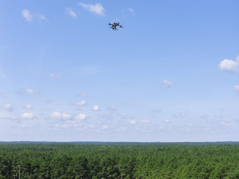 An aerial view of an Ocean County Sheriff's Office DJI M300T drone with a thermal-capable camera payload at the Ocean County Fairgrounds with the New Jersey Pinelands stretching into the distance, September 4, 2026. A propeller plane can be seen below and right of the drone. Albert Yee for NPR.