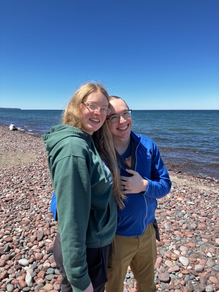 In this photo, Grace Griffin and her husband stand next to one another on the shore of a body of water with many large pebbles along the shoreline. They are smiling for the camera.