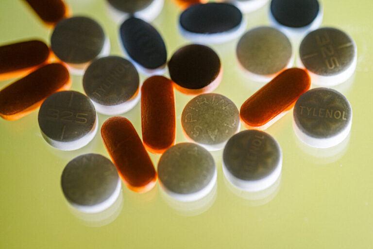 A close-up photo of various over-the-counter pain medicines, including Tylenol, Aspirin, Ibuprofen and Aleve, on a yellow background, hit from behind.