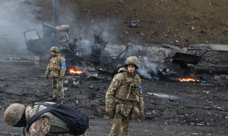 Three Ukrainian service members in camouflage uniforms look for and collect unexploded shells in Kyiv on February 26, 2022. Small fires are behind them among the rubble.
