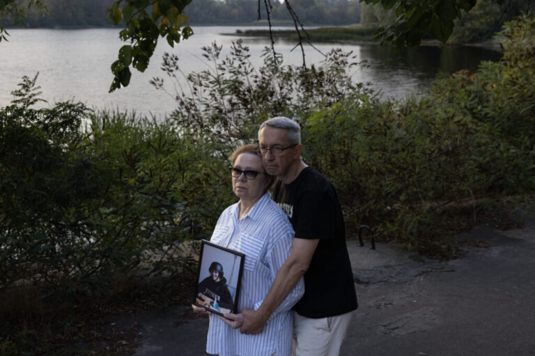 Iryna Mamaysur holds a framed photo of Oleksandra Kuvshynova. Andriy Kuvshynov is embracing her from behind. They are standing by the water in Kyiv.